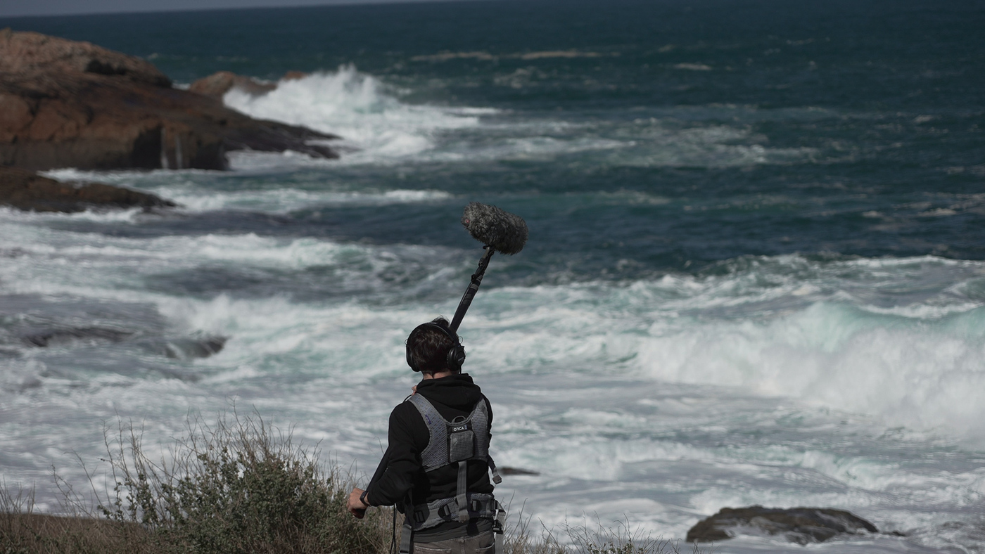 Un home gravando sons cun micrófono de pértiga ao pé do mar